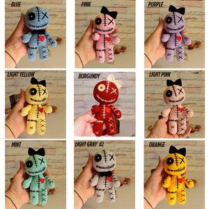 May include: A collection of nine crocheted dolls in various colors including blue, pink, purple, light yellow, burgundy, light pink, mint, light gray, and orange. Each doll features a button eye, an X-stitched eye, a stitched mouth, and a red heart.