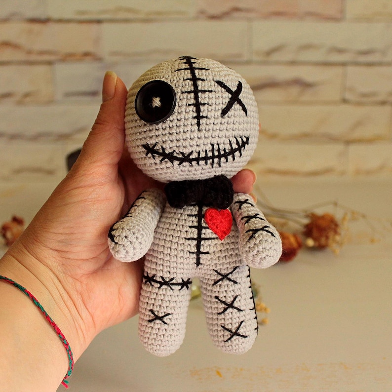 May include: A light gray crocheted doll featuring a black button eye, a stitched X eye, and a wide stitched mouth. The doll wears a black bow tie, has a small red heart on its chest, and includes black stitched X patterns on its limbs and torso.