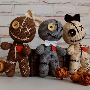 May include: Three handmade crochet dolls in brown, gray, and beige. Each doll features button eyes, stitched mouth details, a small red heart patch on the chest, and a bow on the head or neck. The dolls have visible white or black yarn stitching.