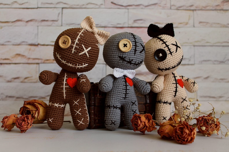 May include: Three handmade crochet dolls in brown, gray, and beige. Each doll features button eyes, stitched mouth details, a small red heart patch on the chest, and a bow on the head or neck. The dolls have visible white or black yarn stitching.