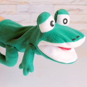 May include: A green frog hand puppet made of soft fleece fabric. The puppet features large white eyes with black pupils, a wide white mouth with a red tongue, and two front legs extending from the body. The design includes stitched nostrils.