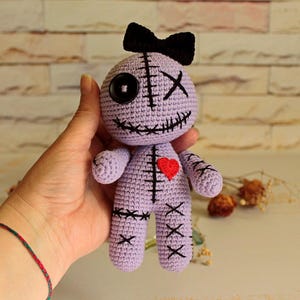 May include: A light purple crochet doll featuring a black bow on its head, one black button eye, and one stitched X eye. The doll has a stitched mouth, a small red heart on its chest, and black stitched X patterns on its arms and legs.