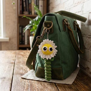 May include: A dark green canvas messenger bag with brown leather accents and a crochet flower keychain attached to the metal hardware. The keychain features a white-petaled flower with a yellow center, large black and white eyes, and a green stem.
