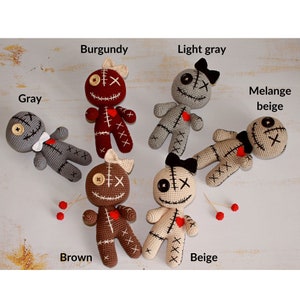 May include: Six crocheted dolls in gray, burgundy, light gray, melange beige, brown, and beige. Each doll features a button eye, an X-stitched eye, a stitched mouth, a red heart patch, and a bow. Text labels include Gray, Burgundy, Light gray, Melange beige, Brown, and Beige.