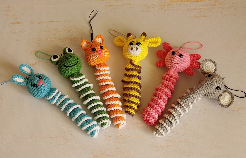 May include: Six crocheted animal toys with long, spiral bodies. The toys are a green frog, an orange cat, a yellow giraffe, a pink axolotl, a gray elephant, and a blue bunny.
