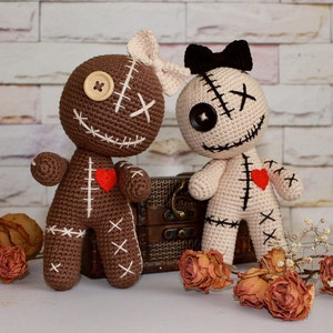 May include: Two crocheted dolls, one brown and one cream, featuring stitched X-shaped eyes, button eyes, and embroidered mouths. The brown doll has a cream bow, while the cream doll has a black bow. Both dolls feature a small red felt heart on the chest.
