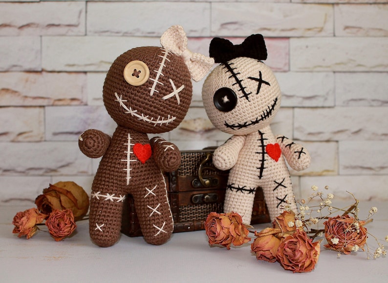 May include: Two crocheted dolls, one brown and one cream, featuring stitched X-shaped eyes, button eyes, and embroidered mouths. The brown doll has a cream bow, while the cream doll has a black bow. Both dolls feature a small red felt heart on the chest.
