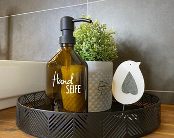 May include: A brown glass soap dispenser with a black pump and a white label that says "Hand Seife". The dispenser is sitting on a black tray with a decorative design. There is a small white bird figurine on the tray.