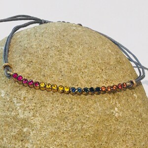 May include: A rainbow colored gemstone bracelet with a gray cord. The bracelet is tied around a gray rock.