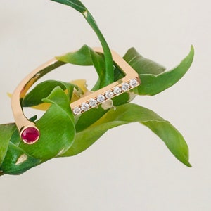 May include: A gold ring with a square shape and a single red stone set in the centre. The ring is adorned with small clear stones along the top edge. The ring is resting on a green bamboo stalk.