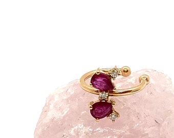 May include: A gold ear cuff with two red gemstones and small clear gemstones. The cuff is displayed on a pink rock.