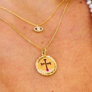 Peut inclure: Un collier en chaîne dorée avec un petit pendentif œil en diamant et un pendentif plus grand avec une croix dorée et une pierre rose.