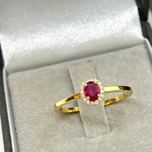 May include: A gold ring with a red oval ruby surrounded by a halo of small diamonds. The ring is in a gray velvet ring box.