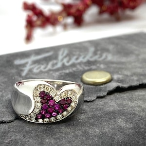 May include: A silver ring with a heart-shaped design set with pink and white gemstones. The ring is displayed on a gray velvet pouch with the text "Fachidis" printed on it.