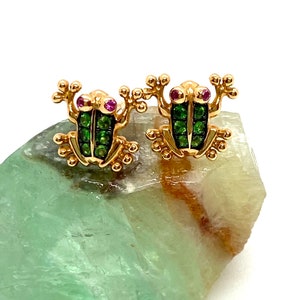 Puede incluir: Un par de pendientes de rana de oro con piedras preciosas verdes y rosas. Los pendientes están sobre una roca verde y marrón.