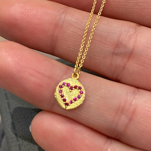 May include: A gold necklace with a small, round pendant featuring a red heart design. The pendant is set with tiny red gemstones.