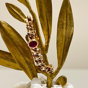 Op de afbeelding: Een gouden kettingarmband met een enkele robijnrode edelsteen. De armband wordt getoond op een gouden tak met bladeren.
