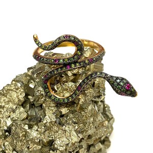 Puede incluir: Un anillo de serpiente de oro con piedras preciosas verdes y rojas. El anillo está sobre un montón de cristales de pirita.