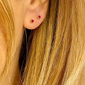 May include: Close-up of a person's ear with two small, round, red gemstone earrings. The earrings are set in gold.
