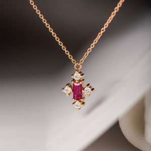 May include: A delicate gold necklace with a pendant featuring a red ruby surrounded by small diamonds. The pendant is shaped like a cross.