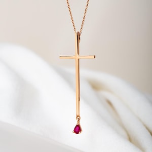 Puede incluir: Un collar con colgante de cruz de oro rosa con una piedra preciosa de rubí en forma de lágrima. La cruz es alargada con una barra vertical y una barra horizontal. El rubí es de color rojo intenso y cuelga de la parte inferior.