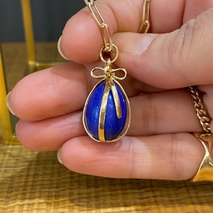 Könnte beinhalten: Eine goldene Halskette mit einem Anhänger in Form eines blauen Lapislazuli-Eies. Das Ei ist in einen goldenen Rahmen gefasst und hat oben eine goldene Schleife.