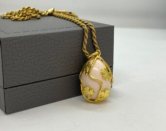 Pode incluir: Um colar de ouro com um pingente de ovo de coral rosa. O ovo é envolto por uma delicada gaiola de ouro com desenhos de folhas.
