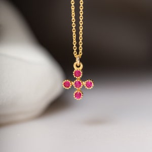 Può includere: Una collana con pendente dorato con un design a croce. La croce è adornata con gemme rosse, rotonde e vivaci. La delicata catena è anch'essa dorata, creando un contrasto sorprendente con le gemme rosse.