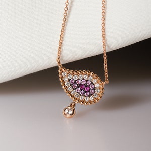 May include: A rose gold necklace with a teardrop-shaped pendant featuring a pink and white eye design. The pendant is surrounded by small beads and has a single diamond hanging from the bottom.