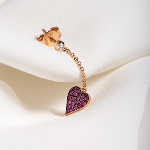 Pode incluir: Um brinco de ouro rosa com um pequeno diamante e um pingente em forma de coração. O coração é coberto por pedras preciosas cor-de-rosa.