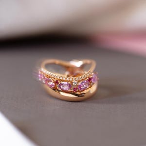 Può includere: Un anello d'oro con un design delicato che presenta una fila di pietre preziose rosa e rosse incastonate in una fascia curva.