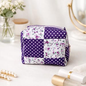 以下が含まれることがあります： パッチワークデザインの小さな正方形の化粧ポーチ。紫色の水玉模様と白い花柄の生地が交互に配置されています。紫色のジッパーと金色の引き手がデザインを完成させています。バッグは白い表面に置かれています。
