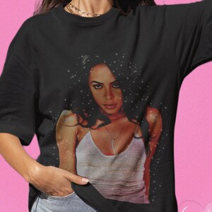 May include: Black t-shirt featuring a graphic print of a woman with long dark hair. The woman is wearing a white vest top and has a sparkling background.