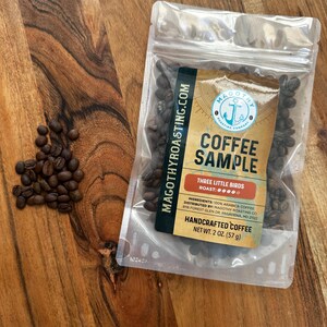 May include: A clear plastic bag filled with coffee beans, labeled "Coffee Sample" with "Three Little Birds Roast." The bag is on a wooden surface, with a small pile of loose beans nearby. The label also says "Handcrafted Coffee" and "Net Wt. 2 oz. (57 g)."
