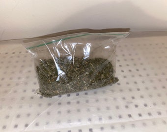 May include: A clear plastic bag filled with dried green herbs. The herbs appear to be a mix of leaves and stems, with a slightly coarse texture. The bag is sealed with a green zipper closure, and the contents are visible.