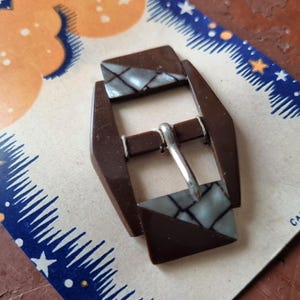 Peut inclure: Boucle marron vintage avec un motif géométrique et un fermoir en métal argenté. La boucle a une incrustation décorative avec un motif bleu clair et gris. La boucle est sur une carte vintage avec un motif bleu et orange.