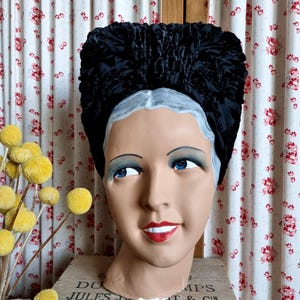 Op de afbeelding: Een zwarte fluwelen tulbandhoed op een mannequinhoofd met blauwe ogen en rode lippenstift. De mannequin staat op een houten kist met de tekst "DU JULES JA... PARIS". De achtergrond is een gordijn met bloemenpatroon.