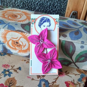 Puede incluir: Dos horquillas para el pelo con forma de flor rosa intenso, con detalles de hilo plateado y una pedrería central. Se presentan en una tarjeta vintage con un retrato de mujer y el texto "MADE IN FRANCE".