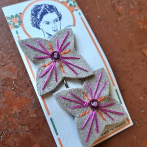 Puede incluir: Dos horquillas hechas a mano con un diseño floral. Las horquillas de fieltro gris presentan bordados rosas y naranjas y una cuenta central púrpura. La tarjeta de respaldo tiene una ilustración vintage y el texto "MADE IN FRANCE".