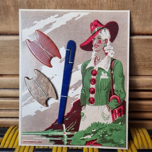 Peut inclure: Une carte postale vintage représentant une femme portant un chapeau rouge et un pull vert, debout dans un paysage montagneux. La carte postale comprend trois aiguilles à tricoter en bois et une aiguille à tricoter bleue.