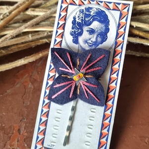 Puede incluir: Horquilla de mariposa de fieltro azul marino con bordado rosa y alfiler de metal plateado. La mariposa tiene un centro en forma de estrella con un acento amarillo y naranja. La horquilla está en una tarjeta vintage.