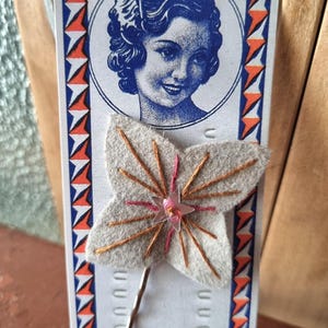 Puede incluir: Una tarjeta de estilo vintage con una ilustración azul y blanca del rostro de una mujer. Una flor de fieltro gris con costuras naranjas y un centro de estrella rosa está adjunta. La tarjeta tiene un borde decorativo con triángulos naranjas y azules.