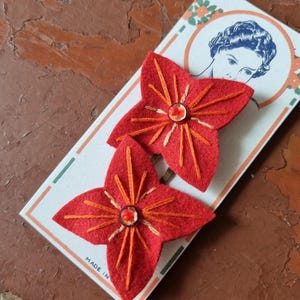 Puede incluir: Dos horquillas para el pelo con forma de flor roja de fieltro, con detalles bordados en naranja y una cuenta central. Se presentan en una tarjeta vintage con un retrato de mujer y el texto "MADE IN".