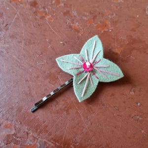 Puede incluir: Una horquilla decorativa con una flor de fieltro verde claro con una gema rosa en el centro y detalles de hilo dorado. La flor está unida a una horquilla de metal plateado, creando un accesorio para el cabello único.