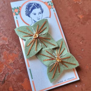 Puede incluir: Dos horquillas de fieltro verde con forma de hoja, con bordados dorados y bronce y una joya central. Se exhiben en una tarjeta vintage con un retrato de mujer y el texto "MADE IN FRANCE".