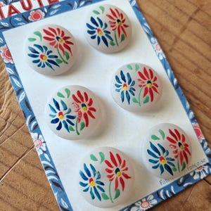 Peut inclure: Six boutons ronds blancs avec des motifs floraux peints à la main en rouge, bleu et jaune. Les boutons sont fixés sur une carte à motifs bleus et blancs avec le texte "HAUTE MODE" en haut.