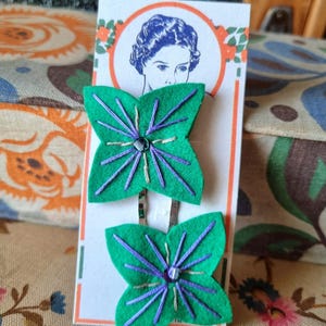 Puede incluir: Dos horquillas para el pelo de fieltro verde con forma de mariposa, con costuras moradas y una cuenta central. Las horquillas se muestran en una tarjeta con una ilustración vintage de un retrato de mujer. El fondo incluye patrones florales.