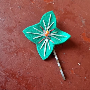 Puede incluir: Una pinza para el pelo de fieltro verde con forma de trébol de cuatro hojas, con detalles bordados en blanco y rojo. El centro presenta una cuenta naranja. La pinza está sujeta a una horquilla metálica plateada.