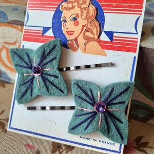 Puede incluir: Un par de horquillas de fieltro verde azulado con forma de flores estilizadas, cada una con costuras moradas oscuras y una gema púrpura central. Las horquillas están sobre una tarjeta vintage con una ilustración retro y el texto "MADE IN FRANCE".