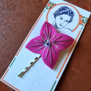 Puede incluir: Una pinza para el pelo con forma de flor de fieltro rosa, con bordados plateados y una joya central. La pinza está sujeta a una tarjeta vintage con un retrato de mujer y el texto "MADE IN FRANCE".